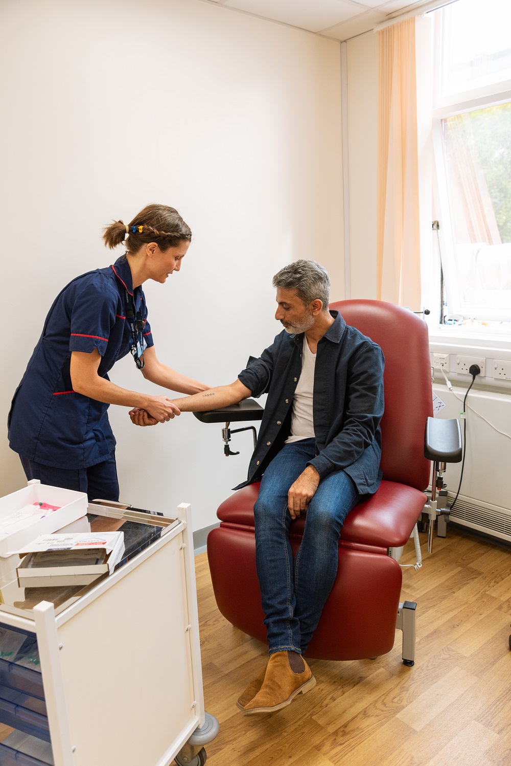 A model takes part in a photoshoot at our Cambridge centre. He is sitting on a red chair holding his arm out to one of our Clinical Nurses