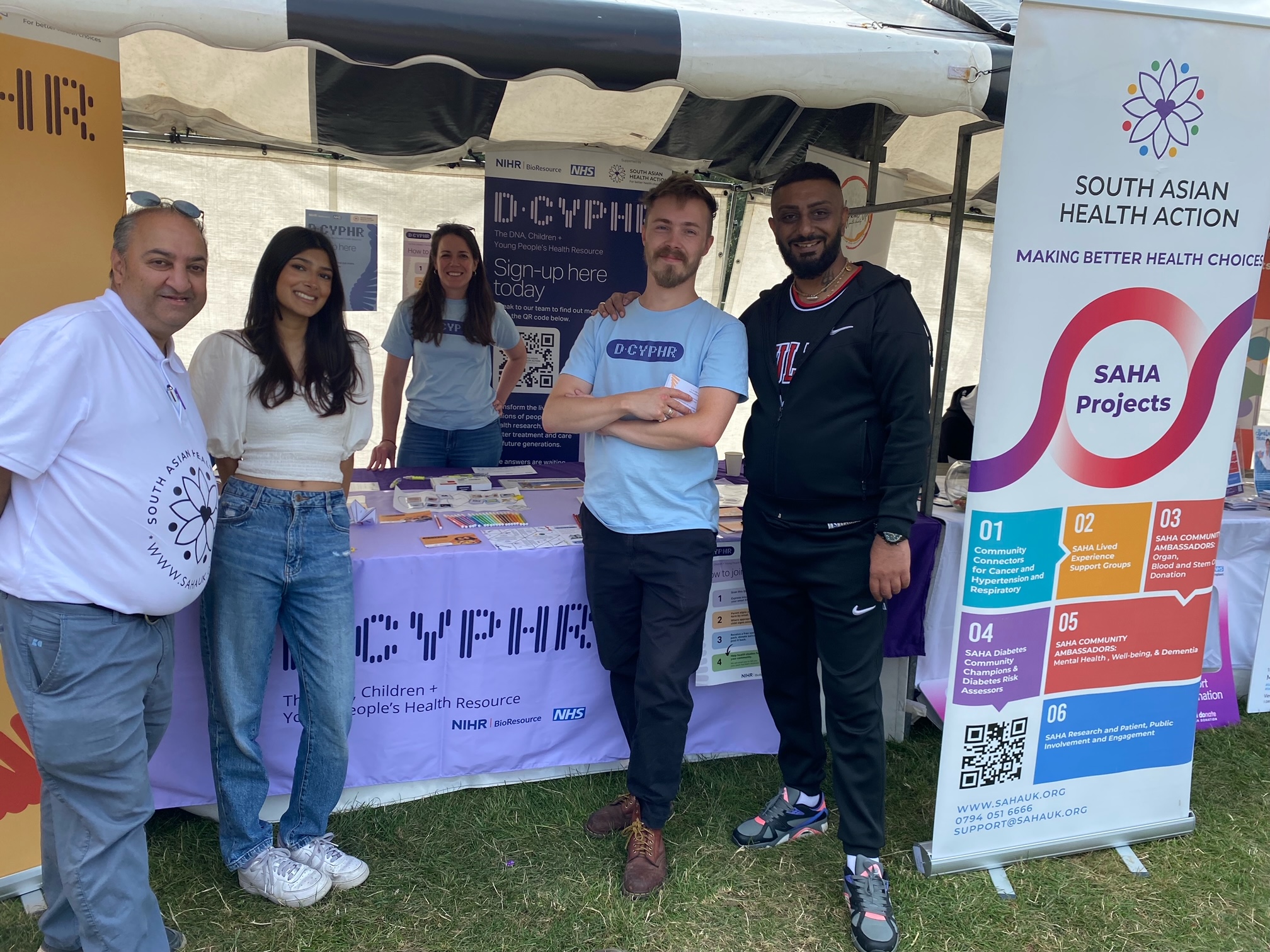 L-R Kirit, Esha, Shayna, James and Keval at the Leicester Mela festival in August 2024. They are standing around a stall that features D-CYPHR banners, promotion posters and leaflets as well as South Asian Health Action content.