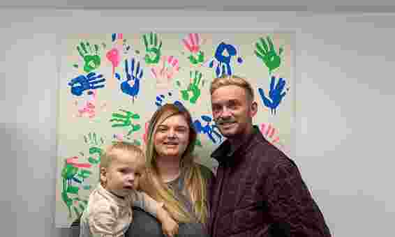 Chloe holding Albert in her arms and stood next to Tom in front of a mural of fluorescent hand prints